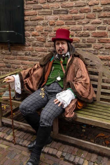 Man in historische kledij met een hoed, zittend op een bankje tegen een bakstenen muur.