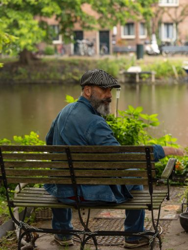 Man met grijs haar en hoed zit op een bankje naast een rustige waterweg.