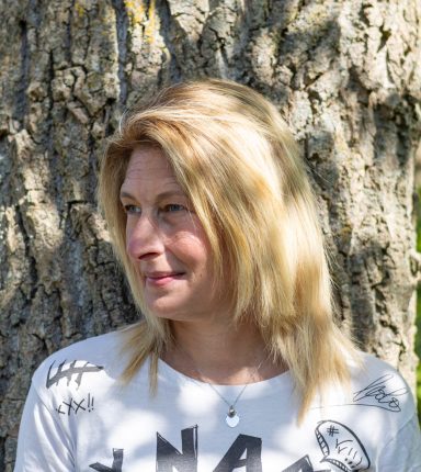 Vrouw met lang blond haar, staand tegen een boom, kijkt opzij.