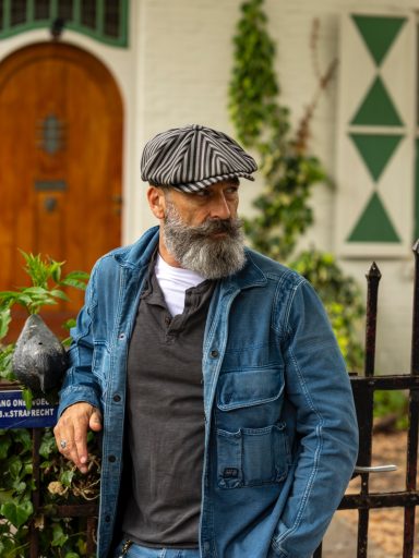 Man met grijze baard en hoed, gekleed in een denimjas, staat bij een hek met planten.