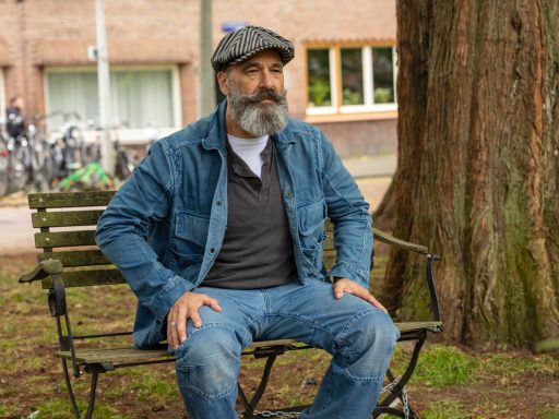 Man met een grijs baard en hoed zittend op een bankje onder een boom.