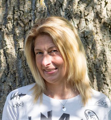 Vrouw met lang blond haar, glimlachend voor een boomstam.
