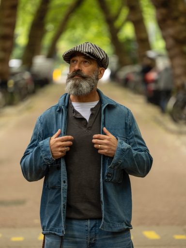 Man met een baard en hoed staat op een straat omringd door bomen en auto's.