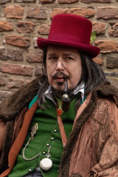 Man in een groen vest met een hoed en een pijp, zittend tegen een stenen muur.