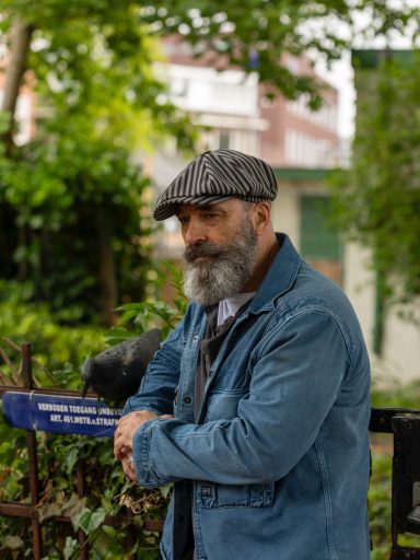 Een oudere man met een grijze baard en hoed staat in een groene omgeving.