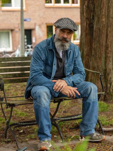 Man met een grijze baard en hoed, zittend op een bank in een park.