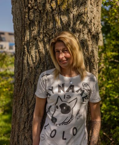 Vrouw met blonde haren, gekleed in een wit t-shirt, leunt tegen een boom.