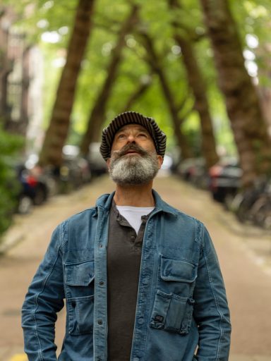 Man met een baard en hoed, kijkend omhoog in een lommerrijke straat.