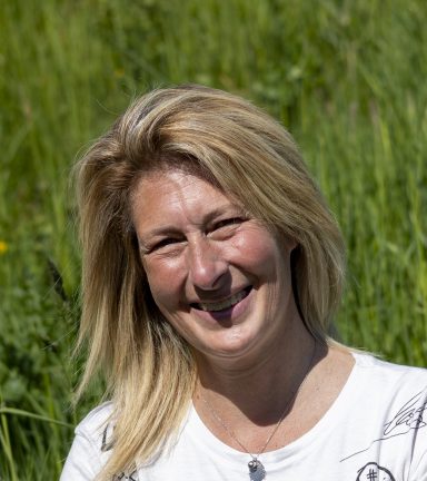 Vrouw met blond haar, glimlachend, staand in een groene, weelderige omgeving.