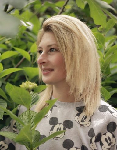Jonge vrouw met blond haar, glimlachend te midden van groene bladeren.