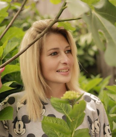 Jonge vrouw met blond haar, omringd door groene bladeren, glimlachend.