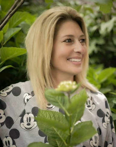 Een vrouw met lang blond haar, staat in een groene omgeving.